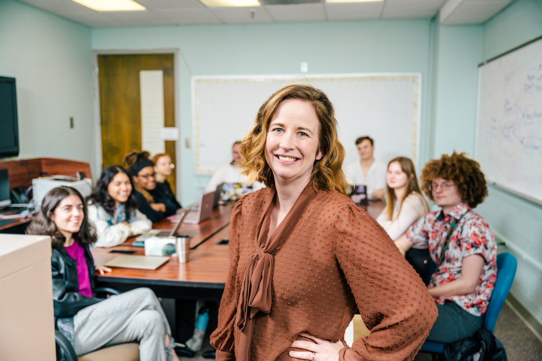 Dr. Michaeline Jensen smiling in a lab meeting room with her students seated around a table behind her